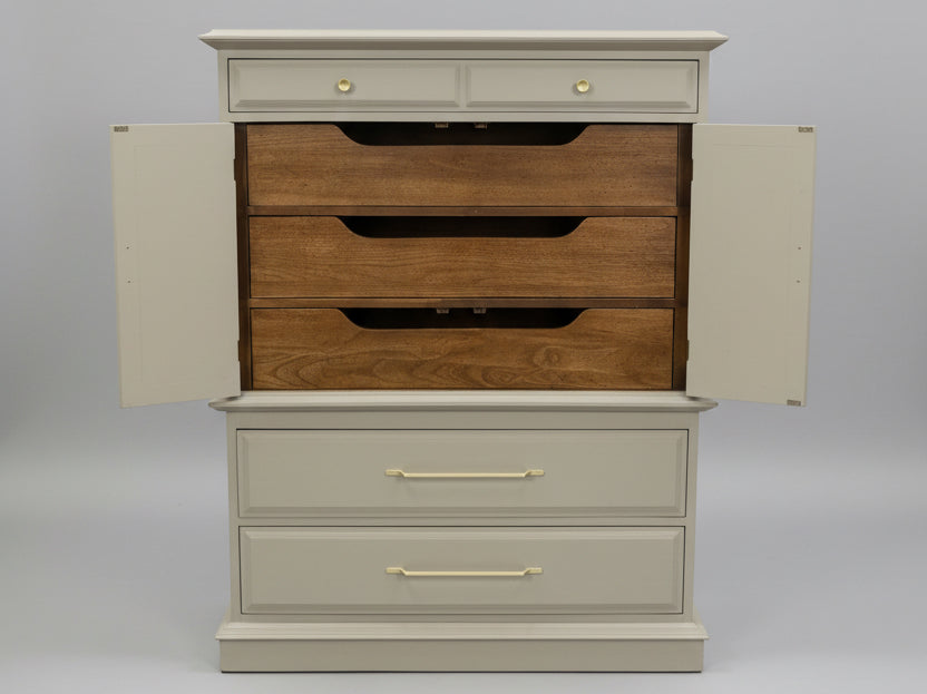 A tall storage chest with a light gray painted finish and multiple drawers, shown in a furniture store. The top two drawers have small round gold knobs, the middle three drawers have dark inlay handles, and the bottom two drawers have longer gold bar handles.